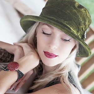 May include: A woman wearing a green velvet hat with a decorative pin. She is wearing a black and red bracelet.
