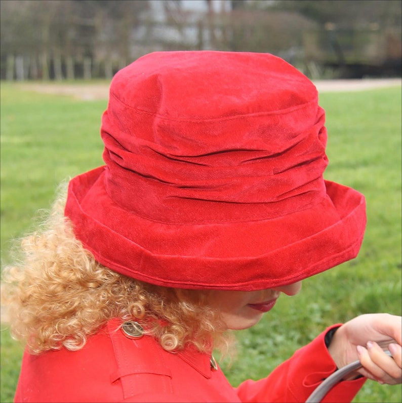 May include: A red velvet hat with a wide brim and a gathered crown. The hat is worn by a person with curly blonde hair.