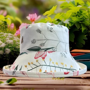 Può includere: Un cappello a secchiello bianco con un motivo floreale con fiori rosa e gialli, foglie verdi e steli grigi. Il cappello ha un'ampia tesa ed è realizzato in tessuto chiaro. Lo sfondo è sfocato con vegetazione.
