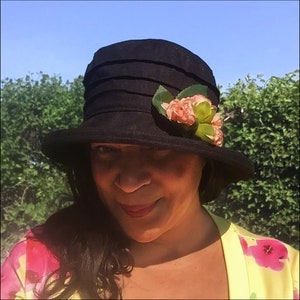 Puede incluir: Un sombrero negro de ala ancha con una corona plisada y un adorno de flor rosa. El sombrero lo lleva una persona con el pelo oscuro y una camisa floral amarilla y rosa.