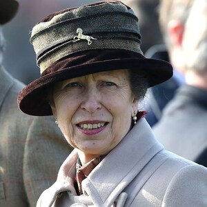 Puede incluir: Una mujer con un sombrero de tweed marr&oacute;n y verde con un alfiler de caballo dorado, un abrigo blanco y pendientes de perlas. Est&aacute; sonriendo.