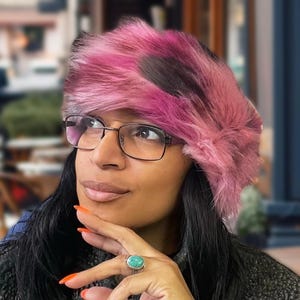 Può includere: Un cappello in finta pelliccia rosa e nera. Il cappello è di un rosa vibrante con accenti neri più scuri. Il cappello è indossato con occhiali e un anello con una pietra turchese.