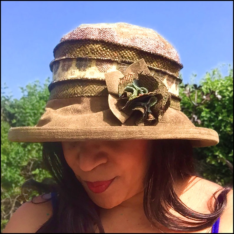 Puede incluir: Un sombrero de fieltro marr&oacute;n con ala ancha y una banda con estampado verde y marr&oacute;n. El sombrero tiene una gran flor de tela verde en un lado.