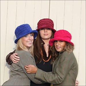 May include: Three women wearing different colored hats. The woman in the middle is wearing a burgundy hat, the woman on the left is wearing a blue hat, and the woman on the right is wearing a pink hat. They are all smiling and looking at the camera.