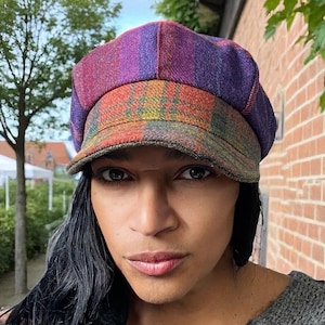 May include: A colorful plaid newsboy cap with a rounded crown and a short brim. The cap features a mix of purple, red, orange, and green hues. The hat is made of a textured fabric.