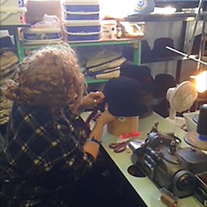Puede incluir: Una persona con una camisa a cuadros est&aacute; trabajando en una m&aacute;quina de coser. Hay varios sombreros en una mesa al lado de la m&aacute;quina.
