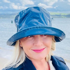 May include: A woman wearing a blue waterproof hat with a wide brim. The hat is made of a shiny, waterproof material. The woman is smiling and looking at the camera.