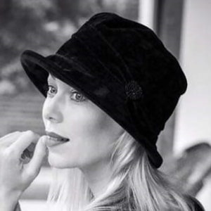 May include: A woman wearing a black velvet hat with a small black embellishment on the side. The hat has a wide brim and is tilted slightly to the side. The woman has long blonde hair and is looking off to the side.