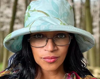 May include: A light blue bucket hat featuring a subtle green leaf pattern. The hat has a wide, downward-sloping brim and a rounded crown. The fabric appears smooth and lightweight, providing full coverage for the head with a soft, flexible structure.