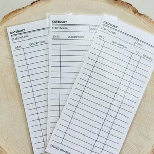 May include: Three white printable budget tracker cards with black text. The cards have headings for category, starting balance, date, description, goal, +/- and balance. The cards are on a wooden surface.