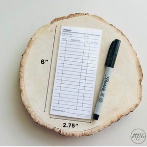 May include: A white card with a grid for tracking finances. The card is 6 inches tall and 2.75 inches wide. The card has headings for category, starting balance, date, description, goal, +/- and balance. The card is on a wooden surface with a black Sharpie pen.