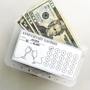 May include: A clear plastic envelope with a white card inside. The card has the text "Anniversary Savings" and a drawing of two wine glasses clinking. The card also has a grid of 25 circles with dollar signs inside. There are several twenty dollar bills inside the envelope.