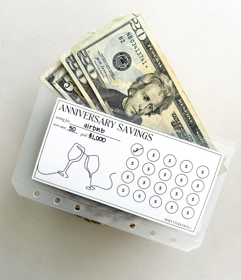 May include: A clear plastic envelope with a white card inside. The card has the text "Anniversary Savings" and a drawing of two wine glasses clinking. The card also has a grid of 25 circles with dollar signs inside. There are several twenty dollar bills inside the envelope.