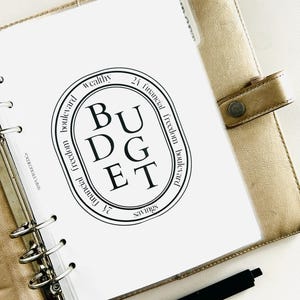 K&ouml;nnte beinhalten: Eine wei&szlig;e Seite in einem goldenen Ordner mit einem schwarzen Kreis, der das Wort "BUDGET" in gro&szlig;en Buchstaben enth&auml;lt. Der Kreis ist umgeben von Text, der "wealthy", "financial freedom boulevard", "24 financial freedom boulevard" und "savings" lautet.