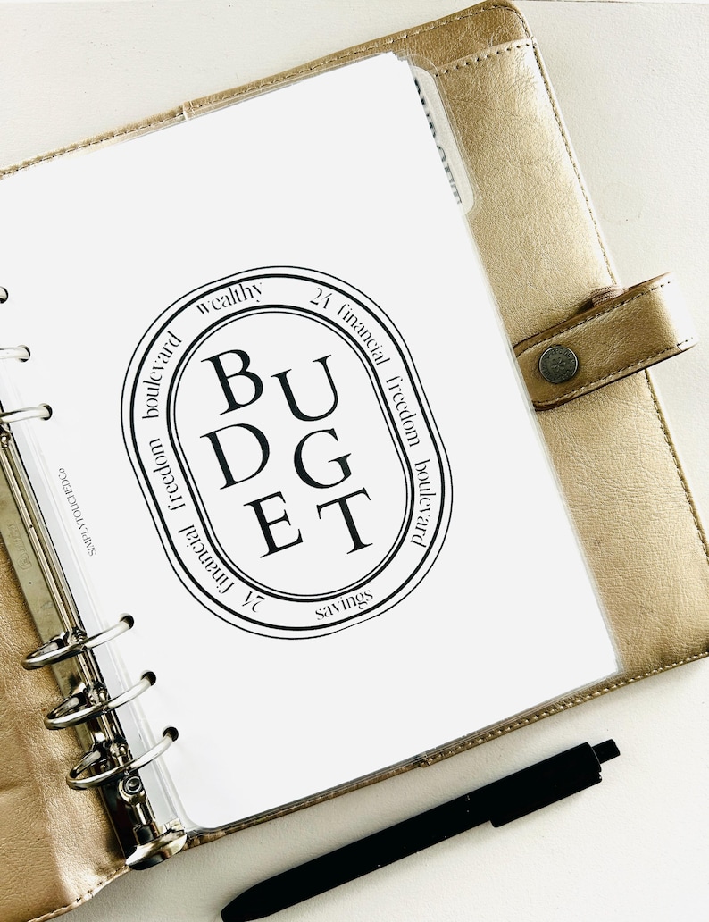 K&ouml;nnte beinhalten: Eine wei&szlig;e Seite in einem goldenen Ordner mit einem schwarzen Kreis, der das Wort "BUDGET" in gro&szlig;en Buchstaben enth&auml;lt. Der Kreis ist umgeben von Text, der "wealthy", "financial freedom boulevard", "24 financial freedom boulevard" und "savings" lautet.