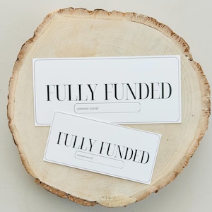 May include: Two white cards with black text that reads "FULLY FUNDED" and a blank line for "amount saved". The cards are on a wooden surface.