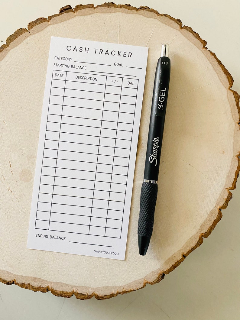 May include: A white cash tracker printable with the words 'CASH TRACKER' at the top. The printable has sections for category, starting balance, goal, date, description, +/- and balance. The printable also has a section for ending balance. The text 'SIMPLYTOUCHEDCO' is at the bottom of the printable. A black pen is next to the printable.