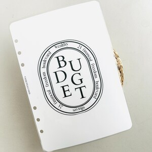 K&ouml;nnte beinhalten: Ein wei&szlig;er Planer mit einem schwarzen Kreis, der das Wort "BUDGET" in Gro&szlig;buchstaben enth&auml;lt. Der Kreis ist umgeben von den W&ouml;rtern "wealthy", "24 financial freedom boulevard", "financial freedom boulevard" und "savings".