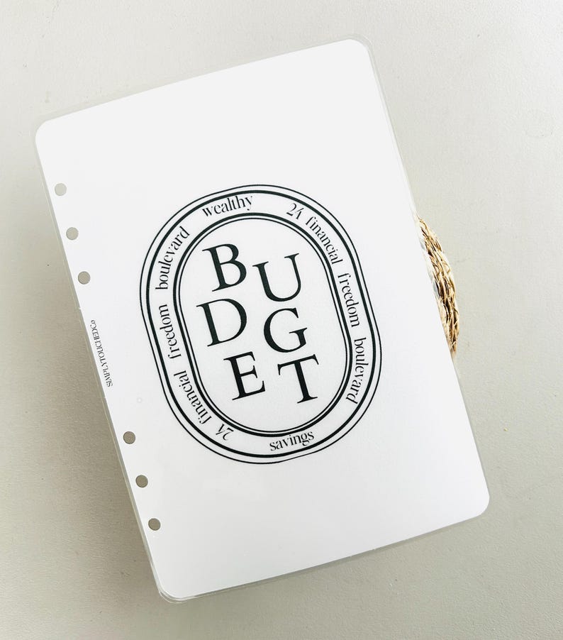 K&ouml;nnte beinhalten: Ein wei&szlig;er Planer mit einem schwarzen Kreis, der das Wort "BUDGET" in Gro&szlig;buchstaben enth&auml;lt. Der Kreis ist umgeben von den W&ouml;rtern "wealthy", "24 financial freedom boulevard", "financial freedom boulevard" und "savings".