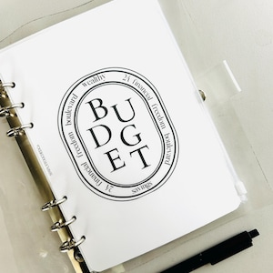 K&ouml;nnte beinhalten: Eine wei&szlig;e Seite in einem durchsichtigen Plastikordner mit einem schwarzen und wei&szlig;en runden Stempel, auf dem "BUDGET" steht, umgeben von den W&ouml;rtern "wealthy", "24 financial freedom boulevard", "savings" und "financial freedom boulevard".