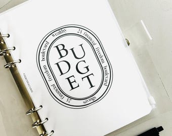 Puede incluir: Una página blanca en una carpeta de plástico transparente con un sello circular en blanco y negro que dice "BUDGET" con las palabras "wealthy", "24 financial freedom boulevard", "savings" y "financial freedom boulevard" alrededor del círculo.