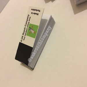 Può includere: Un pacchetto di cartine per sigarette della A UK Roach Company. Il pacchetto è bianco con testo nero che dice "A UK Roach Company Your handiest rolling shop Made in Yorkshire" e un'immagine di un fermaglio per sigarette di cartone animato. Il pacchetto è chiuso e il testo "@aukroachcompany" è stampato sul lato.