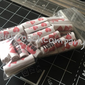 Puede incluir: Una bolsa de plástico transparente llena de pequeños rollos de papel blanco. Cada rollo tiene un corazón rojo y las palabras "I LOVE YOU" impresas.