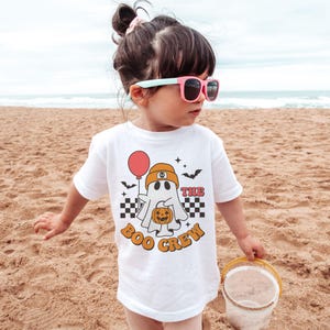 May include: A white t-shirt with a Halloween-themed graphic. The design features a cartoon ghost wearing a beanie and holding a pumpkin and a balloon. The text "THE BOO CREW" is printed in orange. The shirt is worn by a child on a sandy beach.