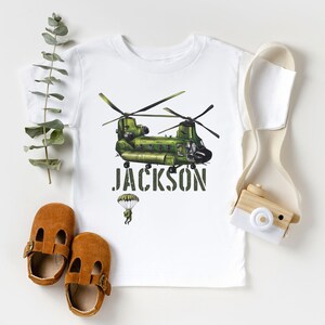 May include: White t-shirt with a green helicopter graphic and the name "JACKSON" printed in black. A small parachute with a figure is below the helicopter.
