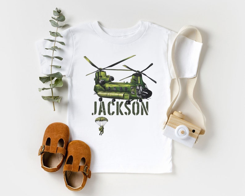 May include: White t-shirt with a green helicopter graphic and the name "JACKSON" printed in black. A small parachute with a figure is below the helicopter.