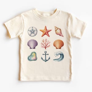 Puede incluir: Una camiseta color crema con ilustraciones de acuarela de vida marina. El diseño incluye una estrella de mar, conchas, coral, un ancla y una ola. La camiseta es de manga corta.