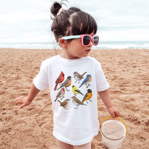 以下が含まれることがあります： さまざまな鳥のカラフルなイラストが描かれた白いTシャツ。砂浜で小さな子供が着ています。鳥には、赤いカージナル、ブルーバード、黄色のフィンチが含まれます。
