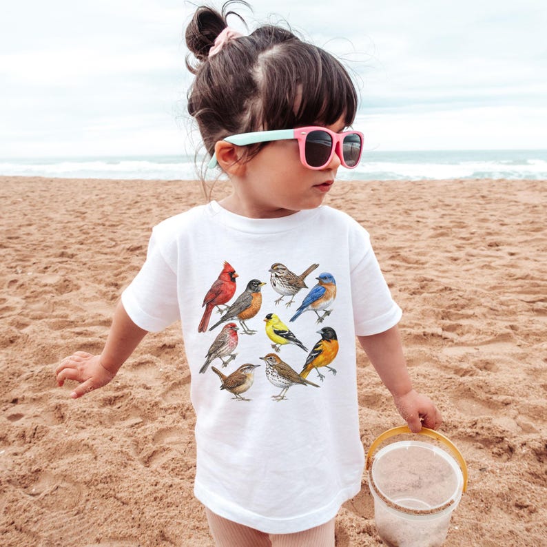 以下が含まれることがあります： さまざまな鳥のカラフルなイラストが描かれた白いTシャツ。砂浜で小さな子供が着ています。鳥には、赤いカージナル、ブルーバード、黄色のフィンチが含まれます。
