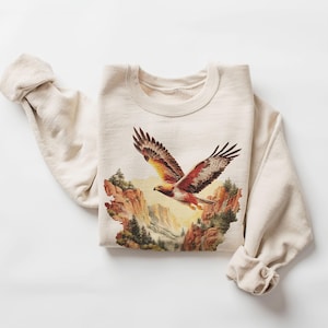 Op de afbeelding: Crèmekleurige sweatshirt met een gedetailleerde afbeelding van een adelaar in vlucht boven een canyonlandschap. De adelaar heeft bruine, witte en gele veren. De sweatshirt heeft lange mouwen met opgerolde manchetten.