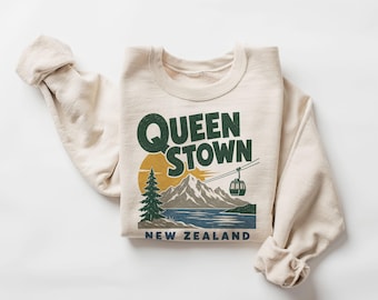 May include: A cream-coloured crewneck sweatshirt featuring a graphic print that reads QUEENSTOWN NEW ZEALAND. The design includes a dark green pine tree, a yellow sun, grey mountains, a blue lake, and a gondola car suspended by thin black lines.
