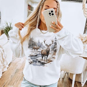 May include: White sweatshirt with a graphic print of a deer in a forest setting. The deer is brown with large antlers.