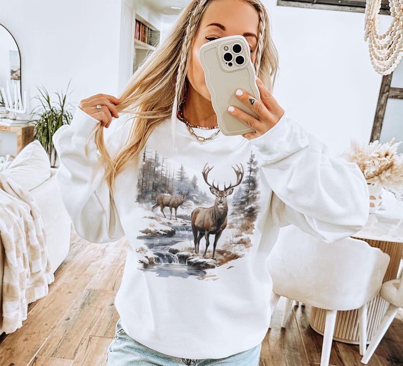 May include: White sweatshirt with a graphic print of a deer in a forest setting. The deer is brown with large antlers.