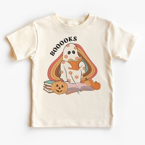 Op de afbeelding: Crèmekleurig T-shirt met een retro design. Het toont een geest die een boek leest, versierd met bloemen, voor een regenboogboog. De tekst "BOOOOKS" staat boven de geest, met pompoenen en boeken eronder.