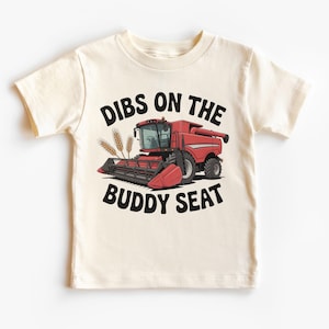 May include: Cream-colored t-shirt featuring a red combine harvester graphic with the text "Dibs on the Buddy Seat" in black. The design includes wheat stalks. A fun, casual tee for kids.