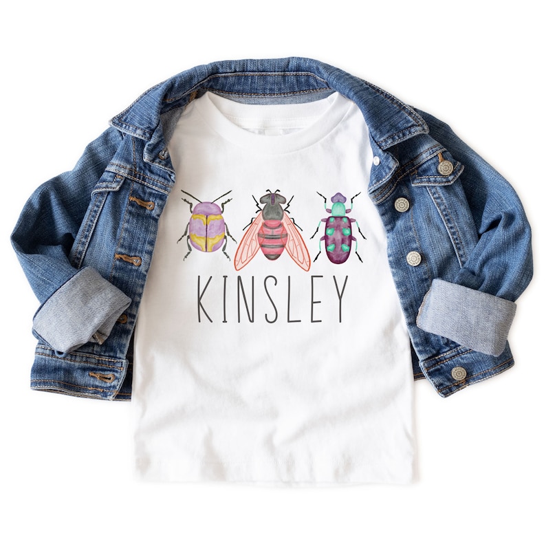 以下が含まれることがあります： 白いTシャツに、水彩画で描かれた3匹の虫と、その下に「Kinsley」という名前が印刷されています。Tシャツは折り畳まれて、デニムジャケットの上に置かれています。