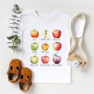 May include: White baby t-shirt with a graphic of nine different types of apples, each with its name printed below. The apples are illustrated in watercolor style. The names of the apples are: Gala, Granny Smith, Honeycrisp, Crispin, Pink Pearl, Fuji, Ginger Gold, Red Delicious, and Black Diamond.