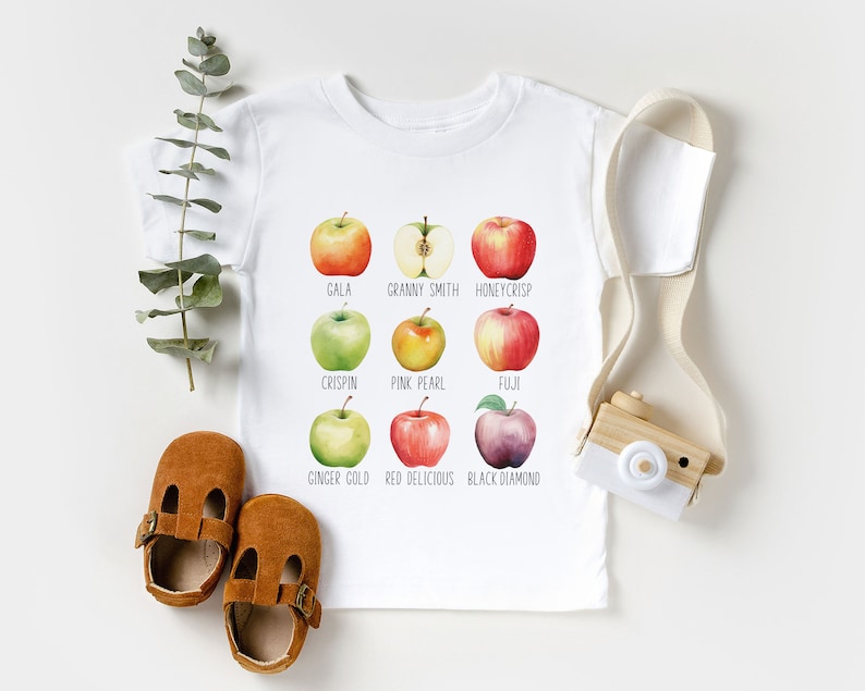 May include: White baby t-shirt with a graphic of nine different types of apples, each with its name printed below. The apples are illustrated in watercolor style. The names of the apples are: Gala, Granny Smith, Honeycrisp, Crispin, Pink Pearl, Fuji, Ginger Gold, Red Delicious, and Black Diamond.