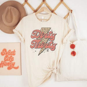May include: A beige t-shirt with a red and black graphic that says "Dirty Thirty" with a leopard print lightning bolt. The shirt is hanging on a wooden rack with a brown hat and a beige tote bag with red sunglasses.
