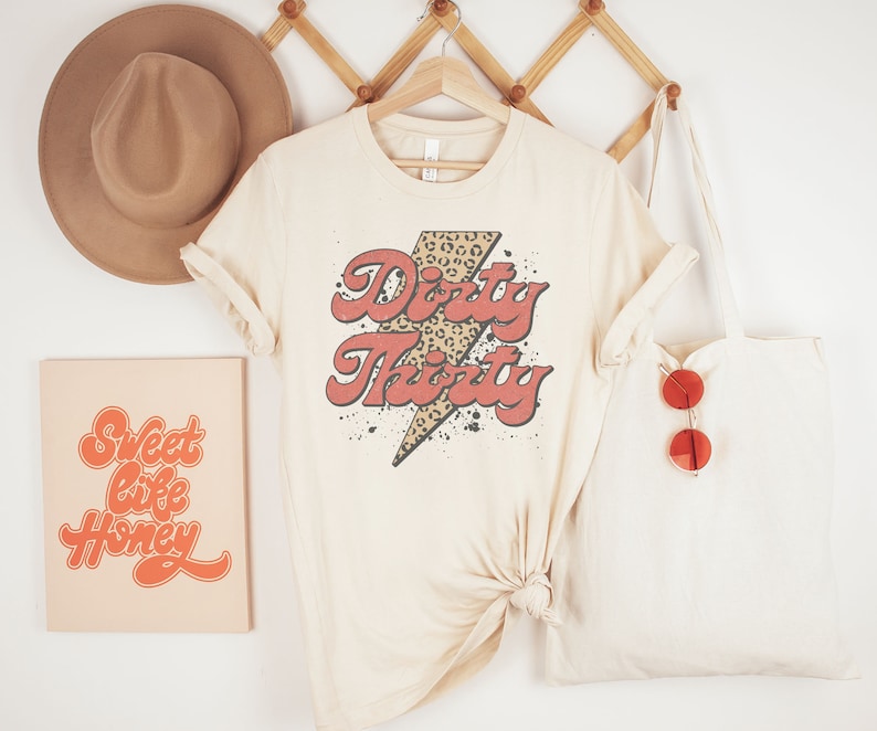 May include: A beige t-shirt with a red and black graphic that says "Dirty Thirty" with a leopard print lightning bolt. The shirt is hanging on a wooden rack with a brown hat and a beige tote bag with red sunglasses.