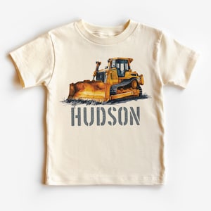May include: A cream colored t-shirt with a yellow bulldozer graphic and the name "HUDSON" printed in gray.