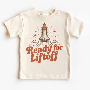 May include: Cream-colored t-shirt featuring a retro-style graphic of a space shuttle launching. The shuttle is orange and gray, with the text "Ready for Liftoff" in orange script below. The design includes stars and clouds.