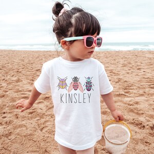 以下が含まれることがあります： 白いTシャツに、カラフルな虫3匹と「Kinsley」という名前がプリントされています。ビーチで子供が着ています。