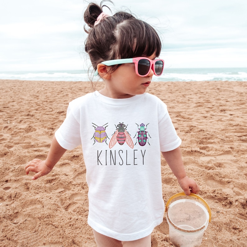 以下が含まれることがあります： 白いTシャツに、カラフルな虫3匹と「Kinsley」という名前がプリントされています。ビーチで子供が着ています。