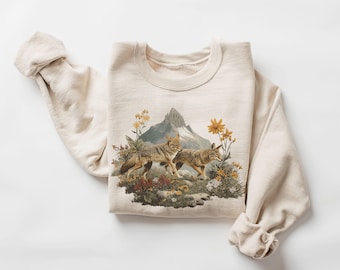 Op de afbeelding: Crèmekleurige sweatshirt met een gedetailleerde afbeelding van twee coyotes te midden van wilde bloemen en een berg op de achtergrond. De top met lange mouwen heeft opgerolde manchetten en een ronde hals.