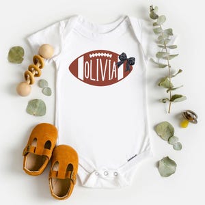 Puede incluir: Body de beb&eacute; blanco con un dise&ntilde;o de bal&oacute;n de f&uacute;tbol americano marr&oacute;n con el nombre "Olivia". Un lazo negro est&aacute; adjunto al costado. La imagen incluye zapatos de beb&eacute;, un juguete de madera y ramitas verdes.